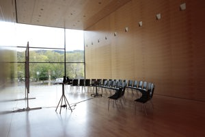 Zweireihig im einem Halbkreis aufgestellte schwarze Stühle vor einer Leinwand im Saal Propter Homines