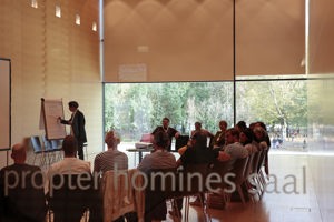 Workshop-Teilnehmer:innen sitzen in einem Halbkreis und eine Person schreibt auf ein Flipchart im Saal Propter Homines