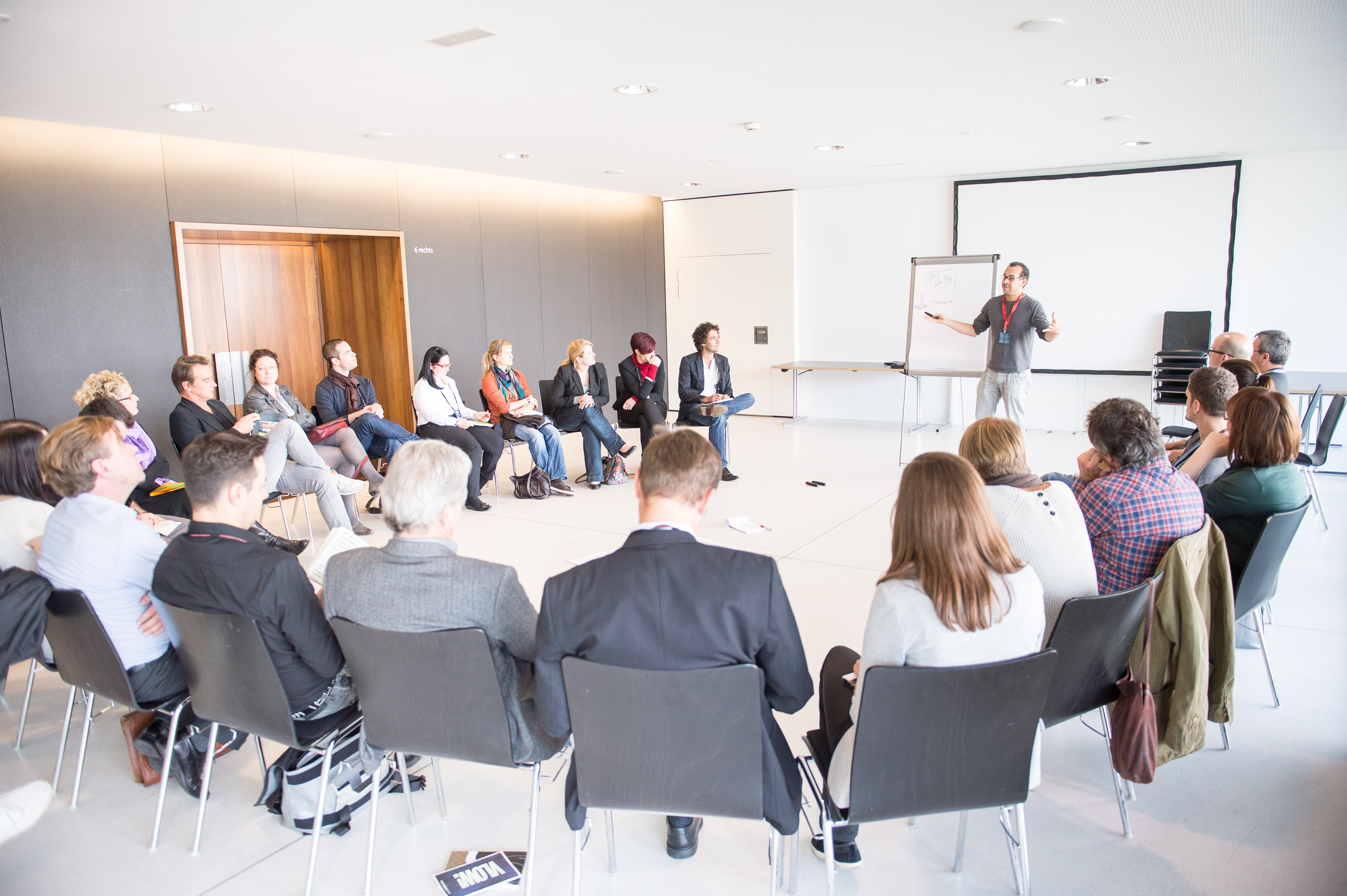 Workshopteilnehmer:innen in einem Stuhlkreis und einem Vortragenden mit Flipchart und Leinwand in der weißen Seegalerie © Matthias Rhomberg