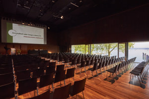Leere Reihenbestuhlung mit schwarzen Stühlen und Mittelgang vor einer Bühne mit Podium im Seestudio im Festspielhaus Bregenz