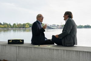 zwei in Anzug gekleidete Männer unterhalten sich am Bodenseeufer, im Hintergrund das Festspielhaus Bregenz
