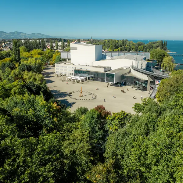 Drohnenaufnahme des Festspielhauses Bregenz inmitten grüner Bäume, im Hintergrund der Bodensee © KoenigsFreunde
